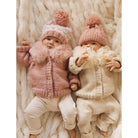 Two babies in pink and beige cardigans and hats lying on a fluffy white surface.