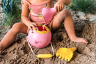 A child sitting in the sand playing with a pink digger toy and a yellow rake toy.