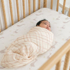 Newborn baby swaddled in a beige blanket lying in a crib with a white sheet.