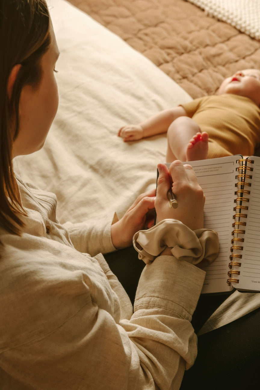 Woman lying on a bed with a baby next to her, holding a notebook.