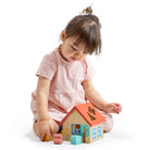 A young toddler in a pink romper sits on a white surface, interacting with a wooden shape sorter house. The child holds a small wooden shape above the roof while several other colorful wooden blocks rest on the floor beside the house.