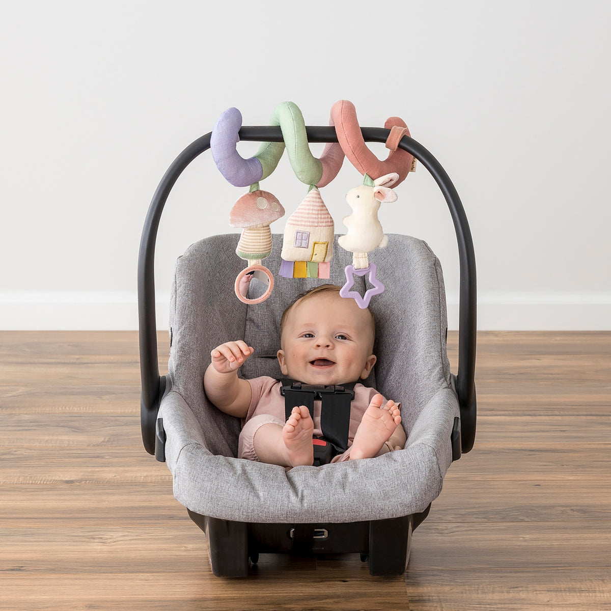 Baby in a car seat with colorful hanging toys on a wooden floor.
