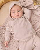 A baby wearing a light beige waffle-knit bonnet and matching long-sleeve romper, lying on a floral muslin blanket in a woven basket.