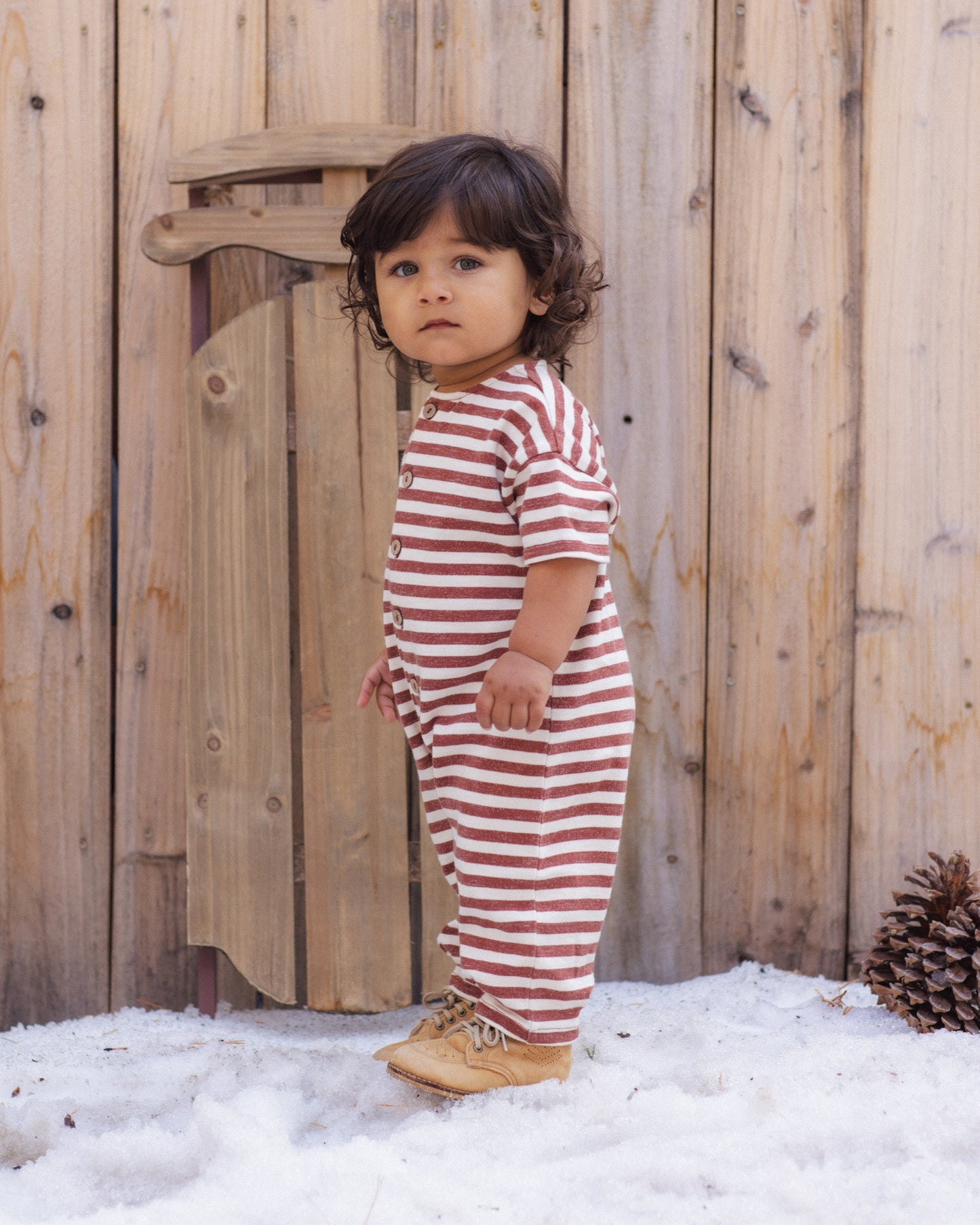 A toddler stands outdoors in front of a wooden fence and snow, wearing a one-piece short-sleeve romper with horizontal red and white stripes and tan boots.