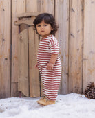 A toddler stands outdoors in front of a wooden fence and snow, wearing a one-piece short-sleeve romper with horizontal red and white stripes and tan boots.