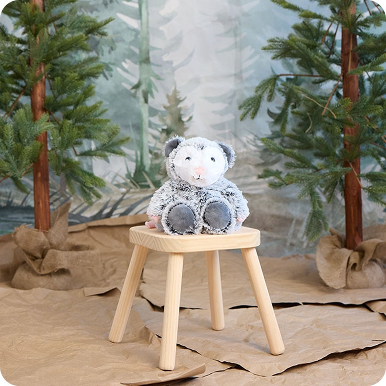 A gray and white fluffy Opossum by Warmies stuffed animal sitting upright on a small wooden stool against a woodland-themed backdrop with trees.