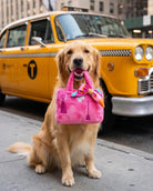 A golden retriever dog sits on a city sidewalk holding a bright pink handbag in its mouth, with a yellow New York City taxi blurred in the background.