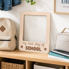 Wooden sign with 'my last day of school' text on a shelf with bags and books.