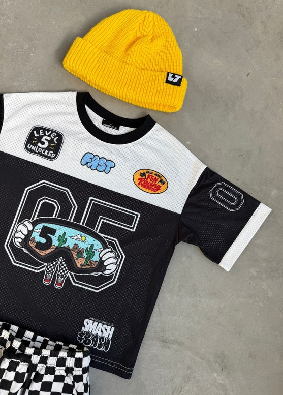 Black and white sports jersey with colorful patches, yellow beanie, and checkered shorts on a gray surface.