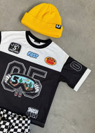 Black and white sports jersey with colorful patches, yellow beanie, and checkered shorts on a gray surface.