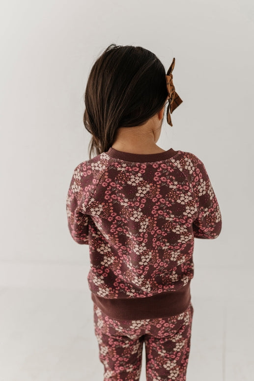 A child viewed from behind wearing a matching long-sleeve top and pants set in a dark burgundy floral print with pink and cream blossoms, against a plain light gray background.