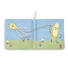 A square fabric book or pouch with a stitched outdoor scene on the cover, featuring a blue sky, green grass, flowers, a yellow kite, and a smiling corn character holding a rope. A white hanging loop is attached at the top center.