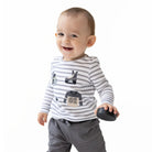A smiling baby boy wearing a long-sleeve gray and white striped shirt with shoe graphics, paired with gray pants, holding a small black toy against a solid white background.