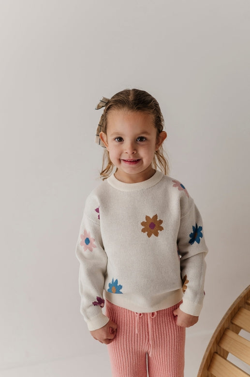 A young child wearing a cream-colored knit sweater with colorful embroidered flowers and pink ribbed pants, standing against a plain light background.