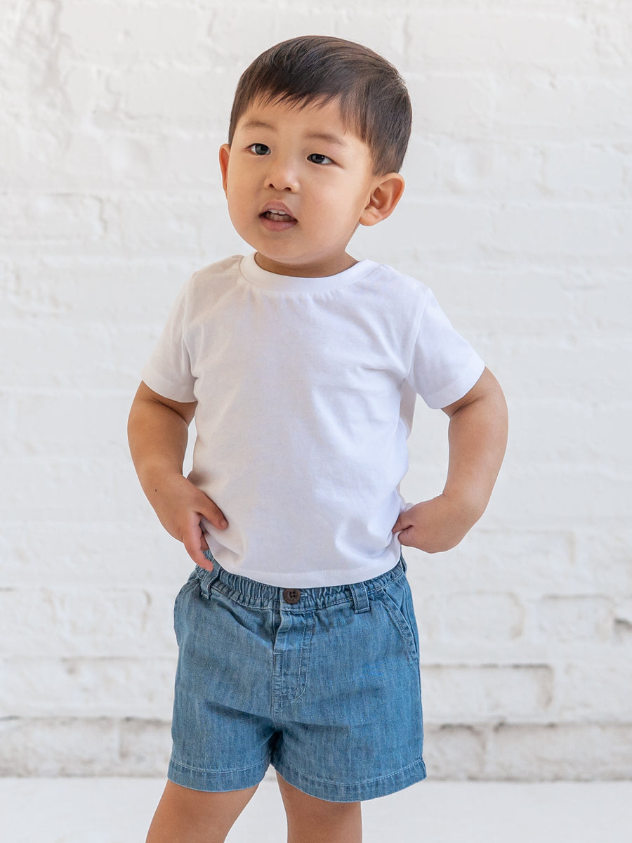 Child wearing a white t-shirt and blue shorts against a light brick wall.