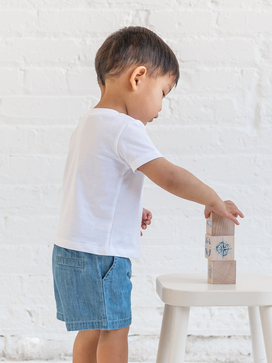 Child in a white t-shirt and blue shorts reaching for a small box on a white stool against a light gray brick wall.