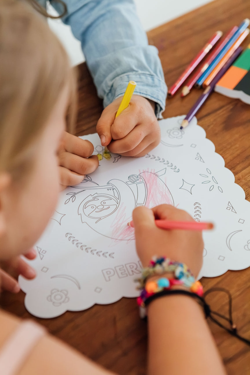 A close-up lifestyle shot of children coloring on a white scalloped paper sheet with a cartoon character. One child is drawing with a yellow pencil, and another is drawing with a pink pencil.