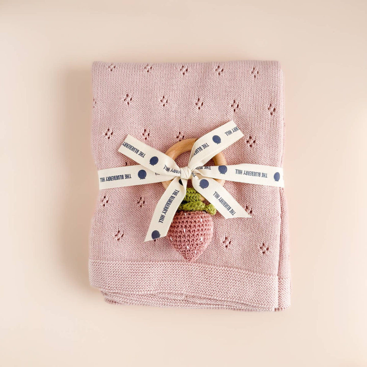 Pink knitted blanket with a wooden spoon and pink strawberry toy, tied with a ribbon on a beige background.
