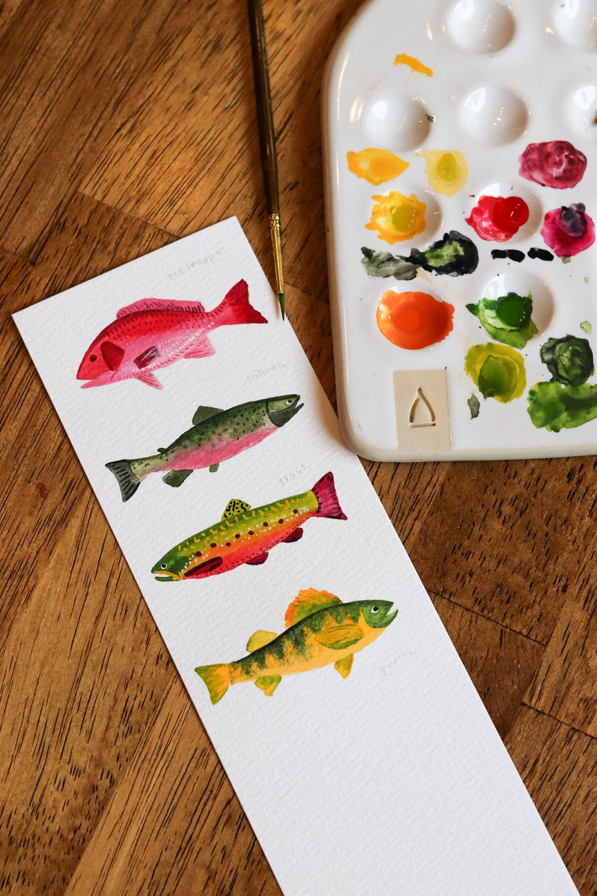 Color swatches with fish illustrations next to a watercolor paint palette on a wooden surface