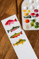 Color swatches with fish illustrations next to a watercolor paint palette on a wooden surface