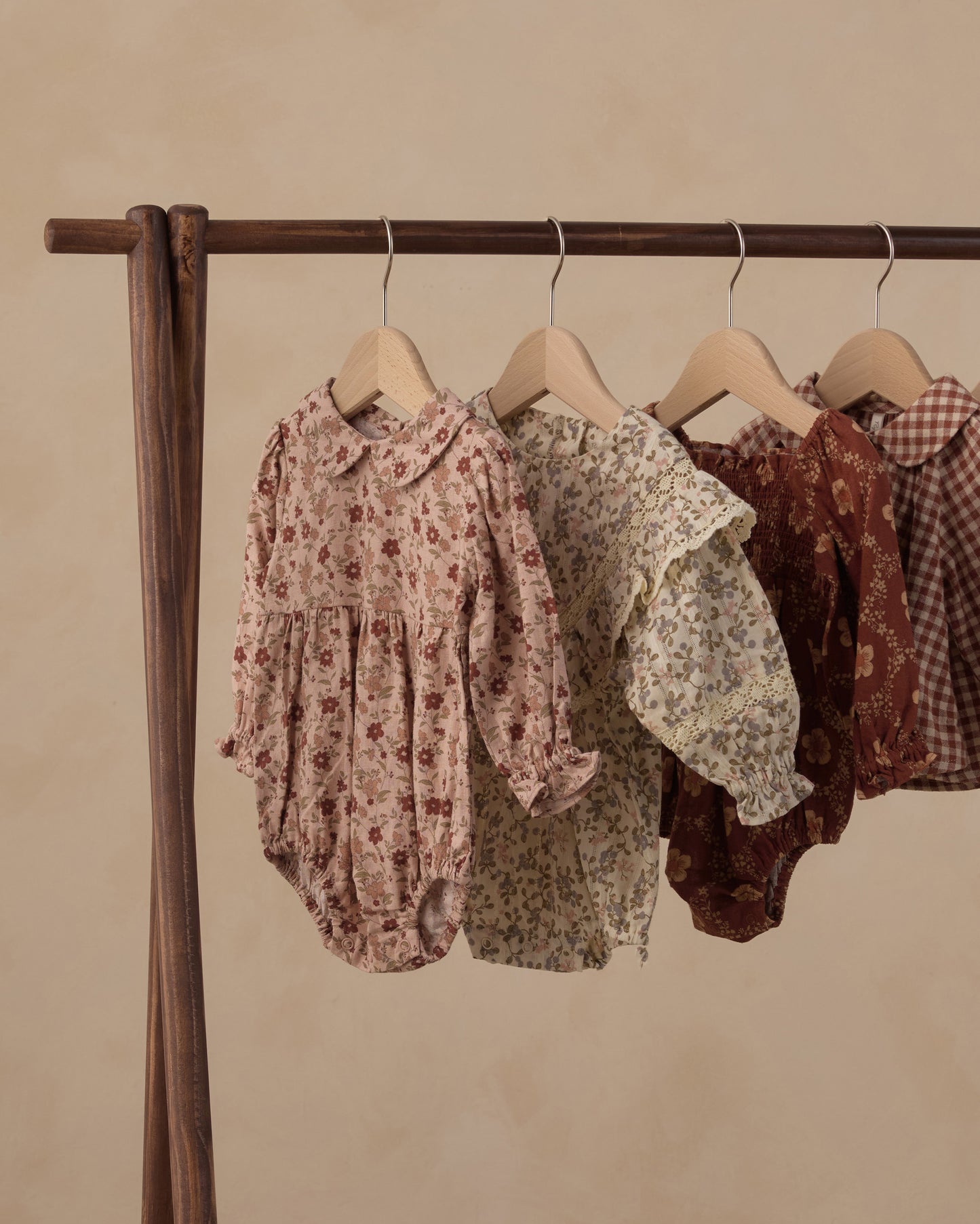 Four baby romper dresses in vintage floral and gingham patterns hanging on wooden hangers from a dark wood clothing rack against a warm beige background.