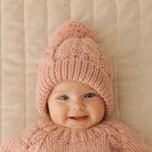 Baby wearing a pink knitted hat and scarf against a beige background