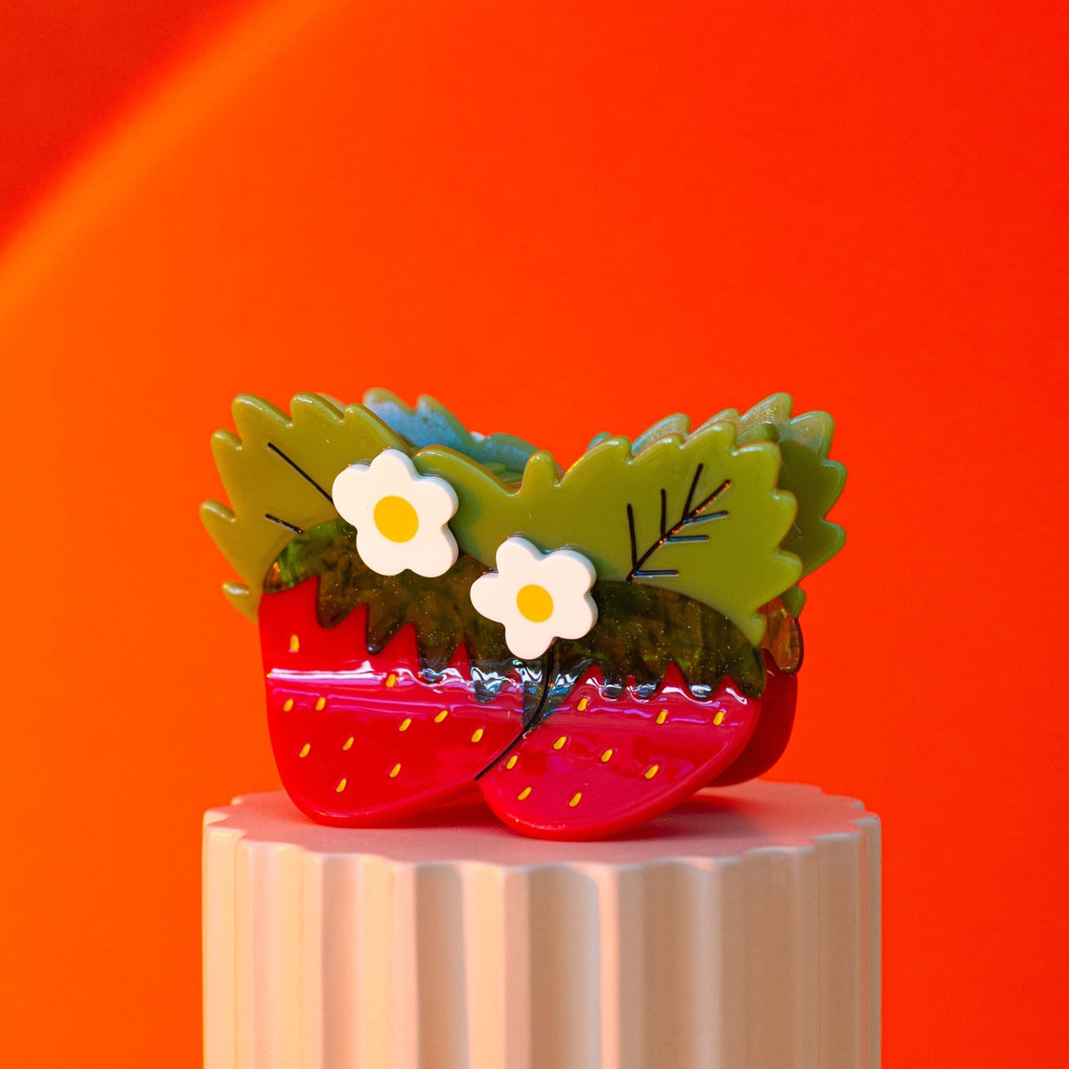 Strawberry-shaped shoes with green leaves and white flowers on a pink pedestal against an orange background