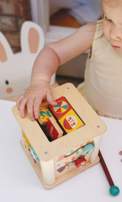 A toddler in a light pink dress interacts with the wooden activity cube by Mentari, reaching out to spin the patterned panels on the side.