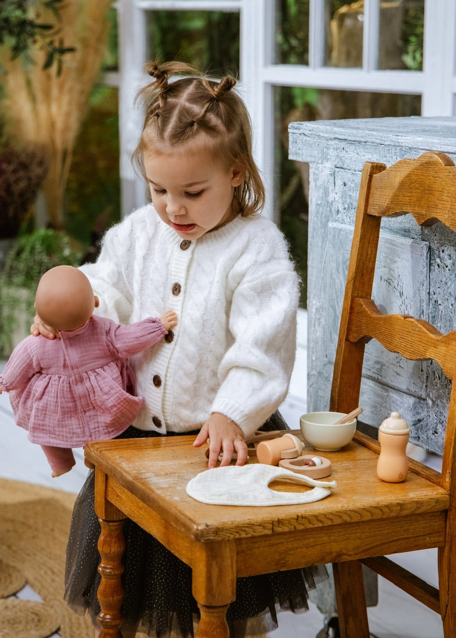 A young girl with braided hair sits on a wooden chair, wearing a white cable-knit cardigan and playing with a pink doll. In front of her on the wooden table are various toy props, including a white bib, a small wooden bottle, and other play items, set against a rustic indoor background with a window and a weathered blue-gray cabinet.