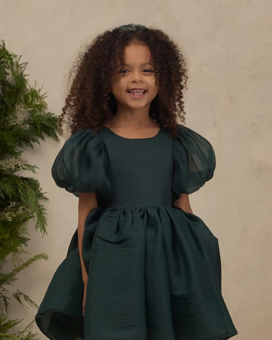 A young girl with curly brown hair wearing a dark forest green dress with puffy sleeves, standing against a textured beige wall with a green plant on the left.