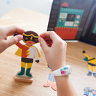 Hands placing a mask onto a magnetic superhero wooden figure from the Superheroes Magnetic Costume Builder set, with other pieces scattered around.