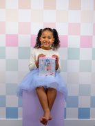 Young girl in a tutu sitting on a purple block against a pastel checkered wall