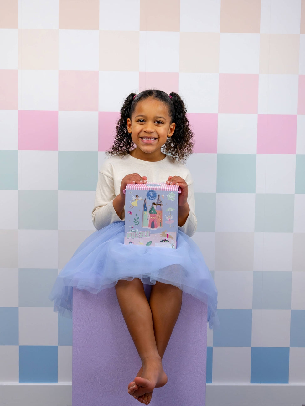 Young girl in a tutu sitting on a purple block against a pastel checkered wall