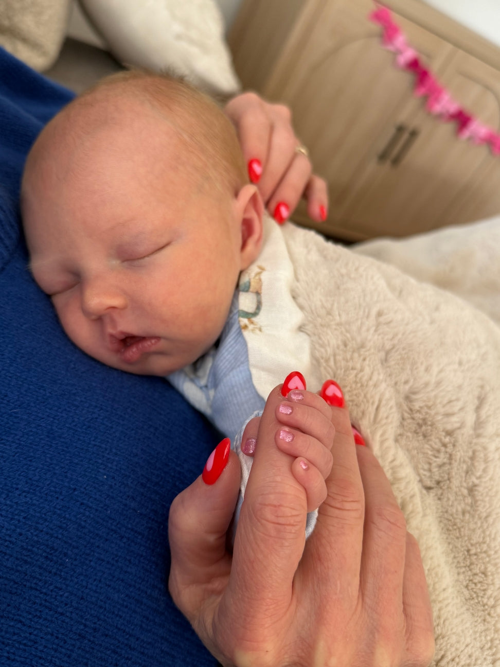 Newborn baby being held by a person with red nail polish, with a blurred background.