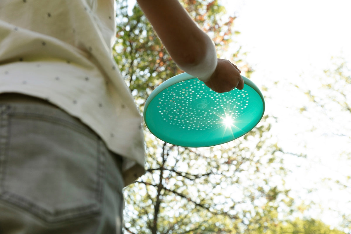 A person holding a bright turquoise flying disc outdoors with sunlight glowing through it.