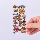Sheet of cartoon bread and pastry stickers with a hand holding the sheet.
