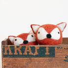 Two small felted wool fox toys in orange and white, nestled inside a rustic wooden crate with a distressed dark green stencil.