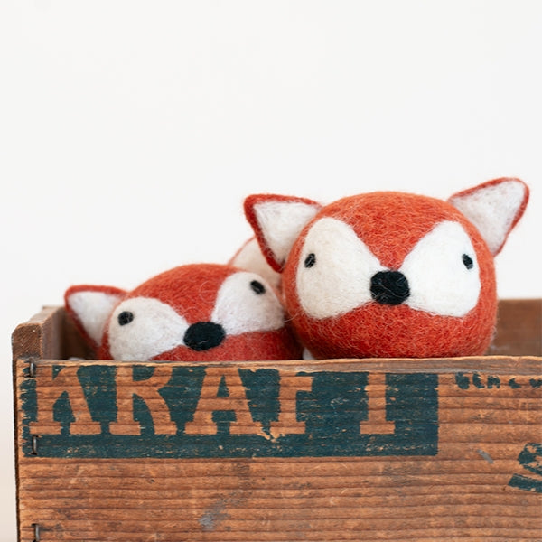 Two small felted wool fox toys in orange and white, nestled inside a rustic wooden crate with a distressed dark green stencil.