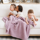 Three children sitting on a couch with a pink blanket