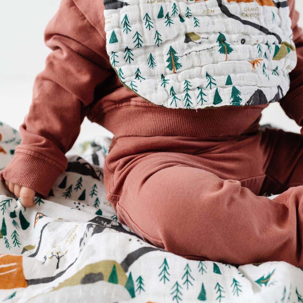 A close-up of a baby wearing a rust-colored sweatshirt and matching pants, sitting on a white quilted blanket with a green forest pattern. The baby is also wearing a matching forest-print bib.