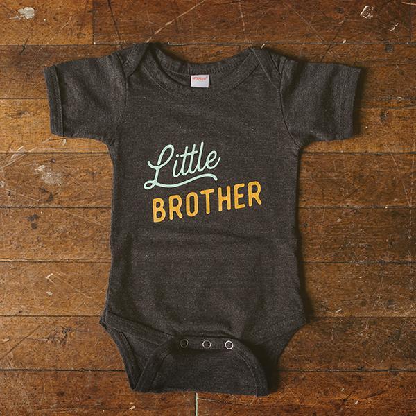 Dark gray baby onesie with 'Little Brother' text on a wooden surface