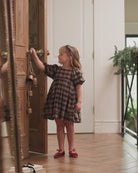 A young girl with blonde hair stands in a doorway wearing a brown and black plaid dress and red shoes, reaching for a brass doorknob on a wooden door.