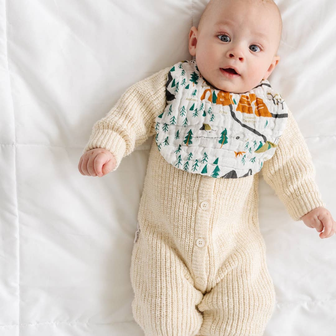 A baby lying on a white quilted blanket, wearing a cream ribbed knit one-piece outfit with a printed bib featuring trees, mountains, and animals.