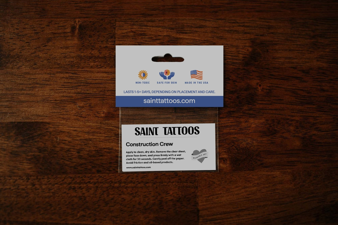 A small rectangular Saint Tattoos card or sticker package labeled 'Construction Crew' photographed from above on a dark wood surface.