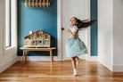 A young child in a light-colored dress skips playfully across a hardwood floor in a brightly lit room, with the Wooden Cedar Chalet Modernist Dollhouse by Mentari resting on a low wooden bench against a deep teal accent wall.