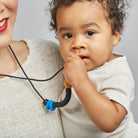 A baby wearing a cream ribbed short-sleeve shirt holds a black silicone teether with a blue and tan accent against their cheek, with a mother's face partially visible on the left.