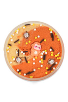 A round, shallow dish filled with orange and beige marbled slime, decorated with small white, yellow, and black sprinkles, chocolate-like pieces, and a central pink bear-shaped topper, photographed on a white background.