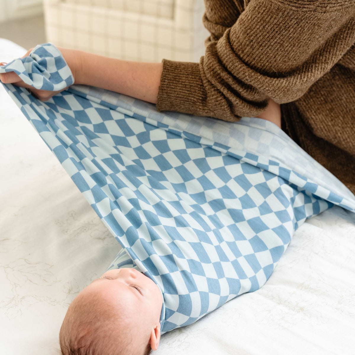 A baby is securely wrapped in a blue and white checkered knit swaddle blanket, with an adult gently holding a matching headband nearby.