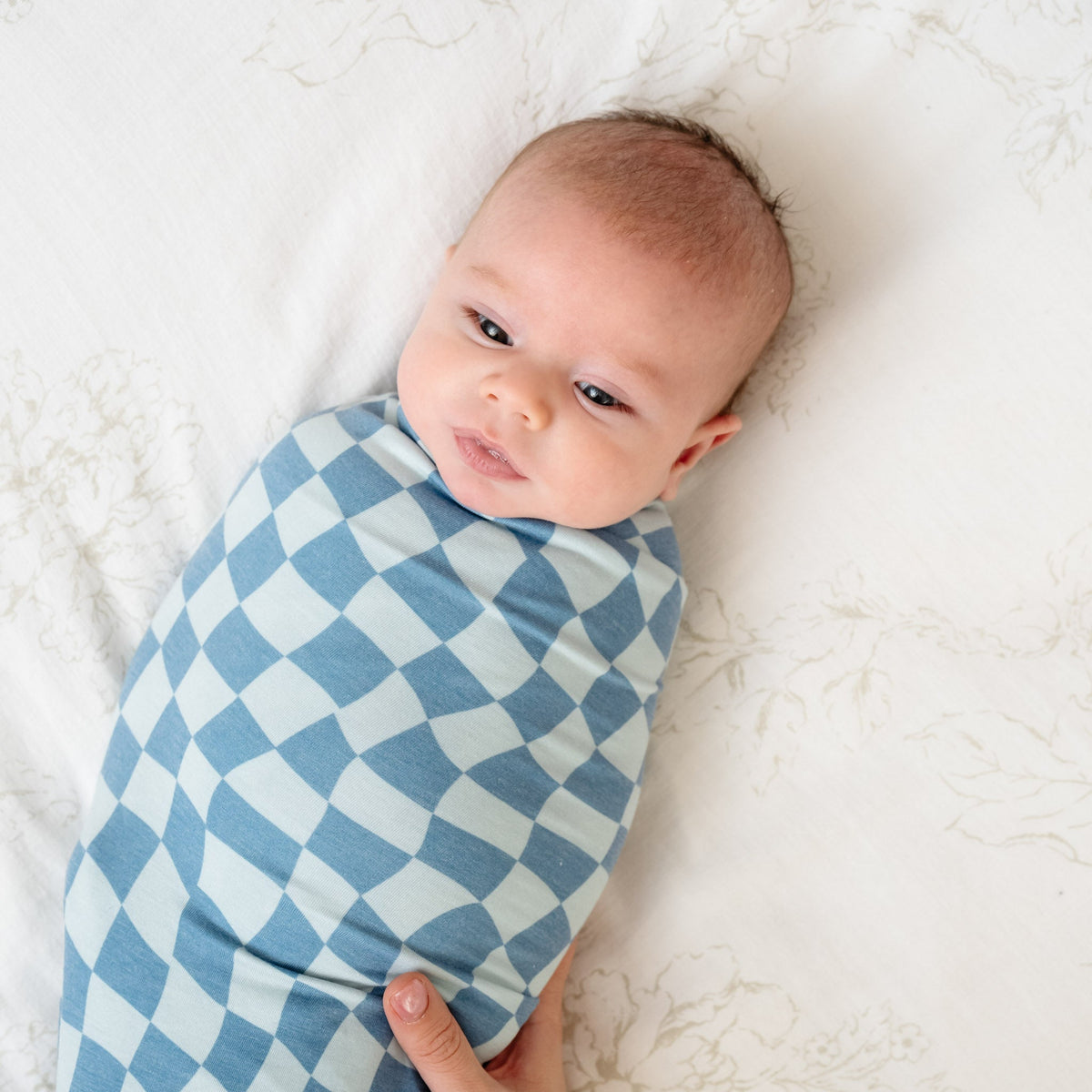 A baby is securely wrapped in a blue and white checkered knit swaddle blanket, with an adult gently holding a matching headband nearby.