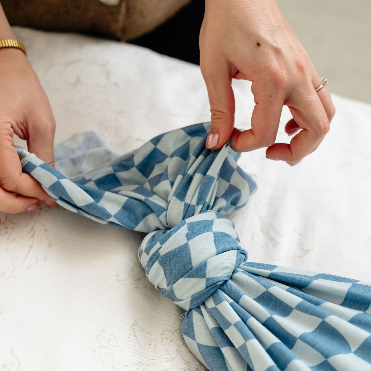 An adult's hands tie a knot in a blue and white checkered knit swaddle blanket against a white background.